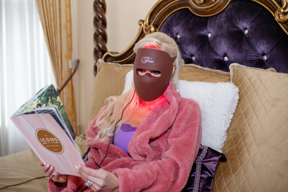 A red light therapy mask with a brownish-gray frame and pink light emitting panels, connected to a controller via a cable.